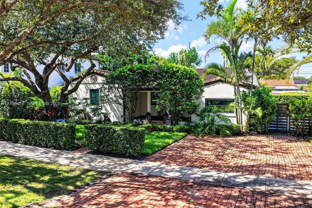 Single-story residence featuring a terracotta tile roof, brick-paved driveway, manicured landscaping, and mature shade trees