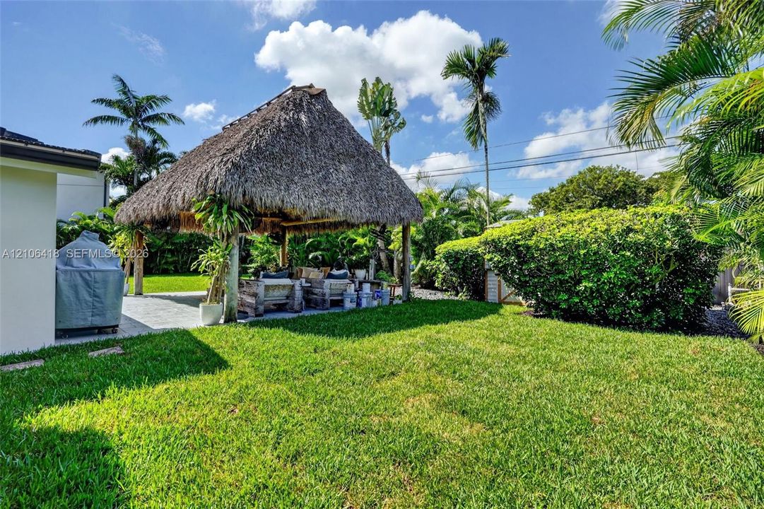 Backyard featuring a thatched-roof tiki hut, paved patio area, manicured lawn, and mature perimeter landscaping