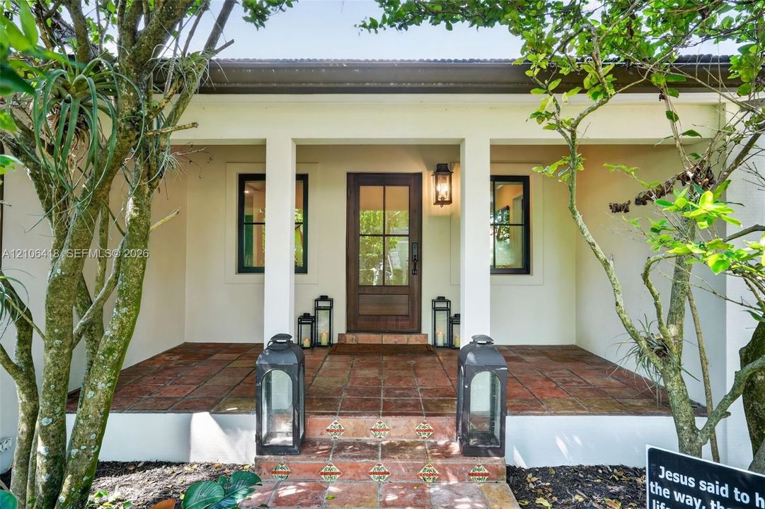 Covered entryway featuring terra cotta tile flooring, decorative riser tiles, a wooden door with glass paneling, and wall-mounted exterior lighting
