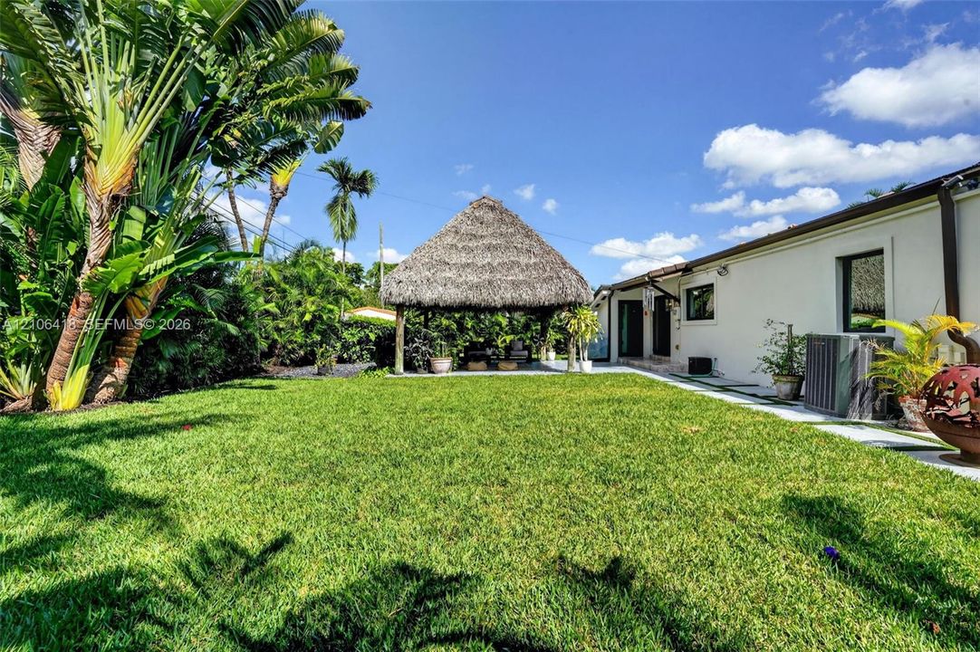 Backyard featuring a thatch-roof gazebo, manicured lawn, established tropical vegetation, and concrete paver walkway