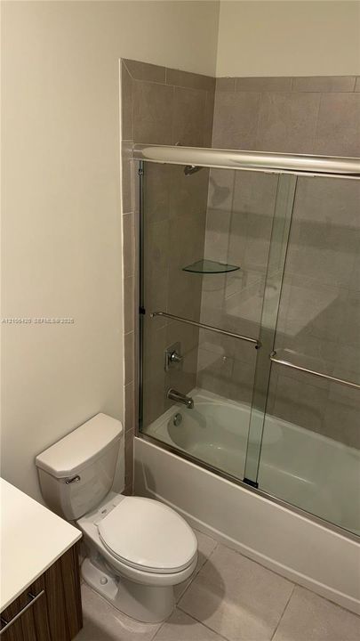 Full bathroom featuring a tub-shower combination with sliding glass doors, neutral tile surround, built-in glass shelving, and a vanity with a solid surface countertop