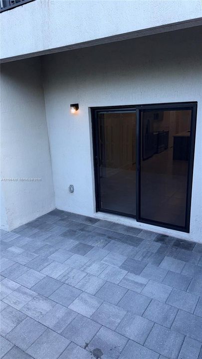 Covered patio featuring gray paver flooring, sliding glass doors with dark frames, and wall-mounted exterior lighting