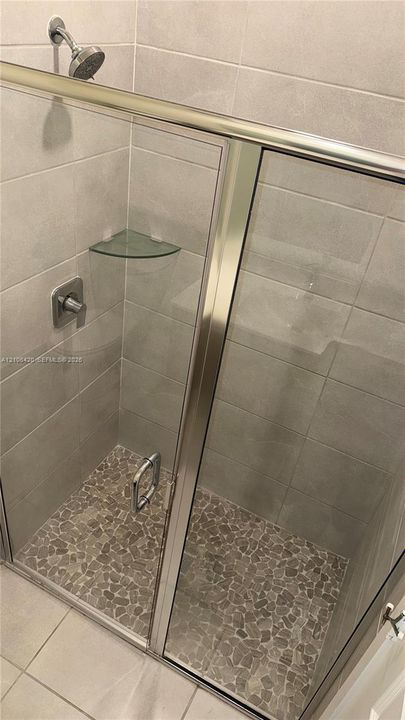 Walk-in shower featuring metallic-finish frame, glass door, pebble-texture stone floor, wall-mounted shower head, and integrated glass corner shelf