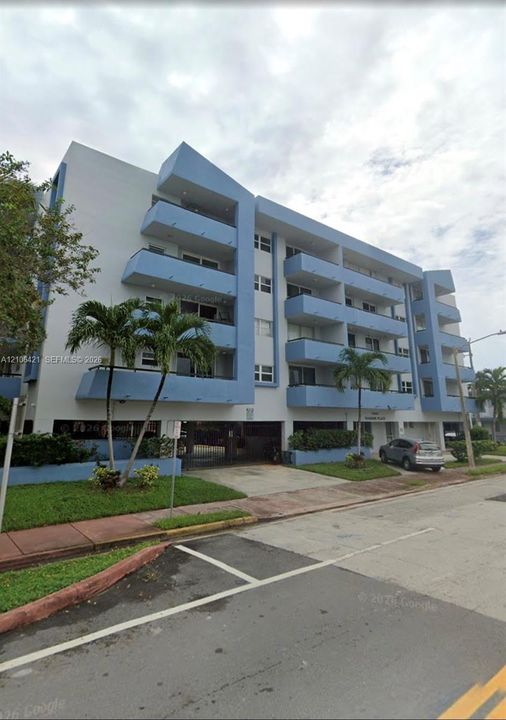 Multi-story residential building featuring blue-accented exterior, private balconies, covered ground-level parking, and landscaped grounds