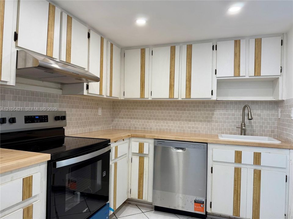 Kitchen featuring custom two-tone cabinetry with wood-finish vertical detailing, butcher-block countertops, a mosaic tile backsplash, stainless steel appliances, and a built-in stainless steel range hood