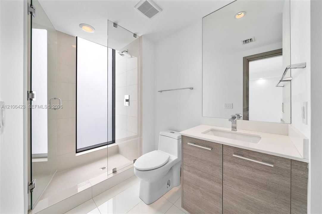 Full-height frameless glass shower enclosure with integrated window, wood-finish vanity with stone countertop, rectangular undermount sink, and polished chrome fixtures