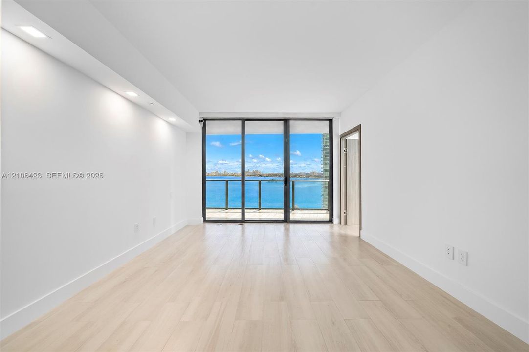 Interior space featuring light-toned wood-finish flooring, recessed ceiling lighting, and floor-to-ceiling glass sliding doors opening to an exterior balcony with water frontage