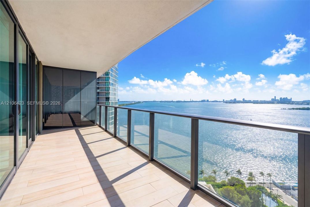 Covered balcony featuring wood-finish tile flooring, floor-to-ceiling glass sliding doors, and a glass panel railing overlooking the bay and skyline