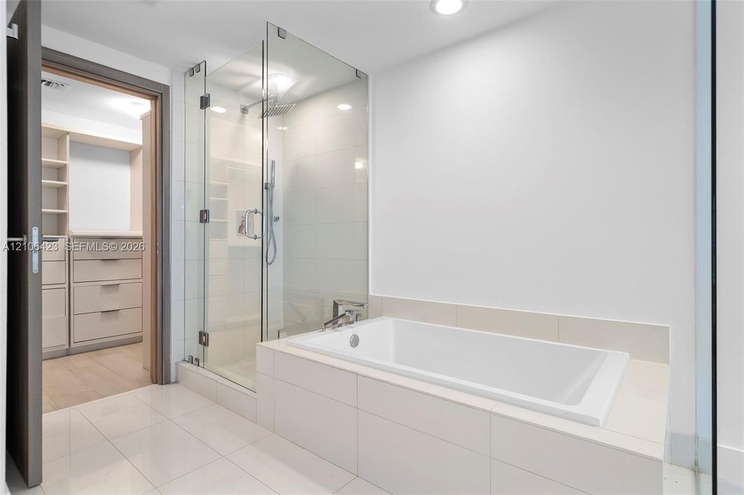 Glass-enclosed walk-in shower with rain showerhead, deep-soaking bathtub with tile surround, built-in custom closet cabinetry, and large-format floor tiling