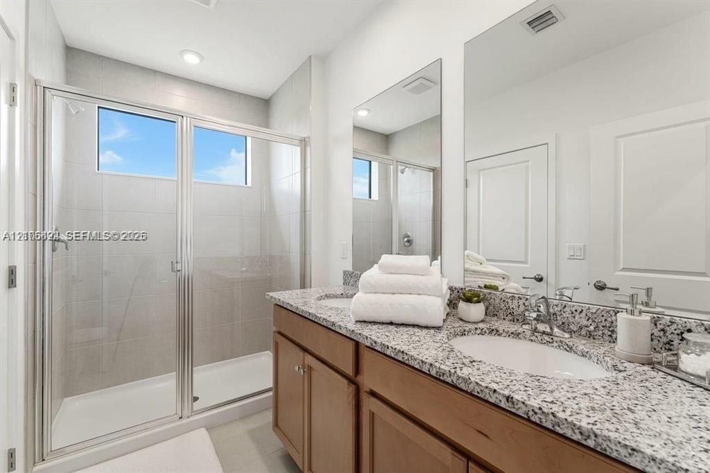 Bathroom featuring a dual-sink vanity with stone-finish countertops, light wood-finish cabinetry, a glass-enclosed shower with tiled walls, and oversized wall-mounted mirrors