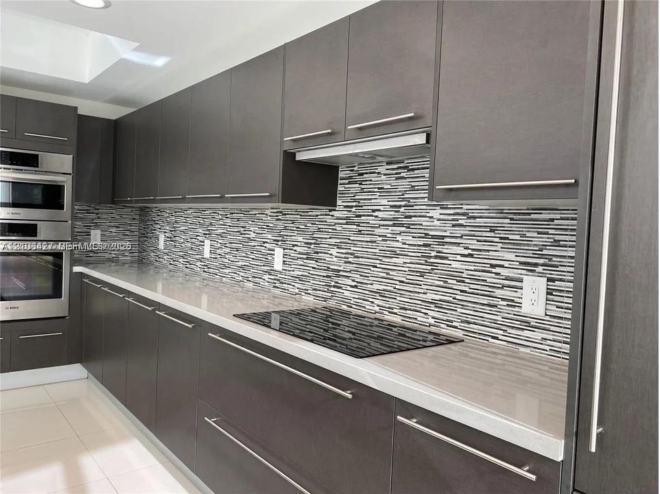 Kitchen featuring flat-panel dark wood-finish cabinetry, quartz countertops, a multi-tonal horizontal mosaic tile backsplash, integrated glass cooktop, and a stainless steel built-in wall oven