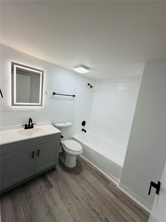 Bathroom featuring a gray shaker-style vanity with a solid surface countertop, matte black fixtures, an integrated LED-backlit mirror, and wood-finish flooring