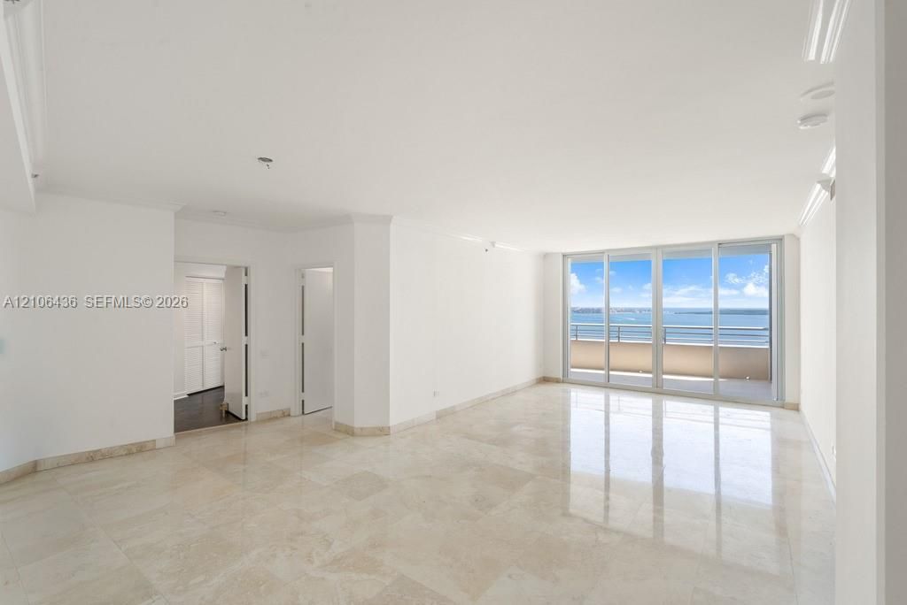 Open-concept interior featuring light-toned stone tile flooring, crown molding, and floor-to-ceiling glass sliding doors providing water scenery and access to a balcony