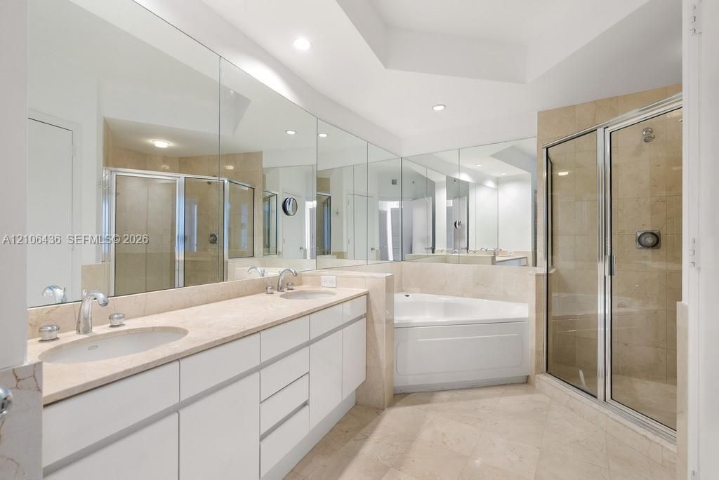 Double vanity with light-toned stone countertop, expansive wall-to-wall mirrors, recessed lighting, corner soaking tub, and enclosed shower with stone-tile surround