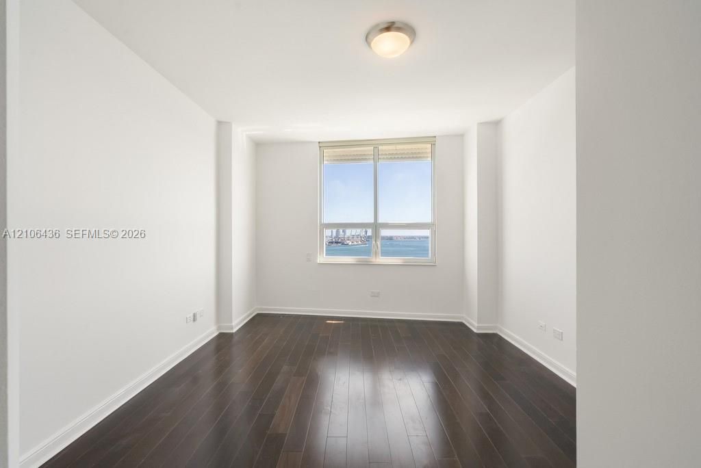 Room featuring wood-finish flooring, white walls, and a large window showcasing water and harbor scenery, complemented by a flush-mount ceiling light fixture
