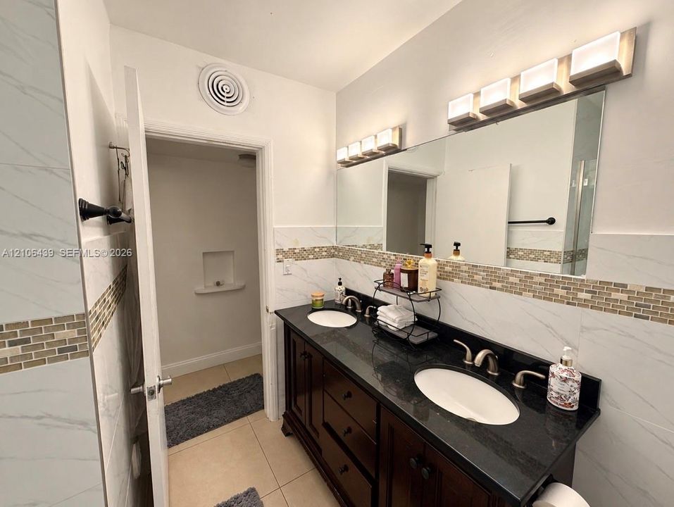 Double-sink vanity with dark cabinetry, stone-finish countertop, white oval undermount sinks, tile-accented backsplash, and dual multi-bulb lighting fixtures