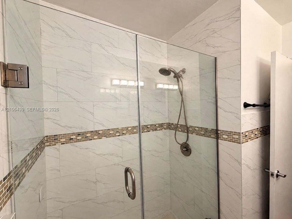 Walk-in shower featuring large-format stone-patterned tile, a glass enclosure with a metallic handle, a decorative mosaic tile accent band, and a multi-setting handheld shower fixture