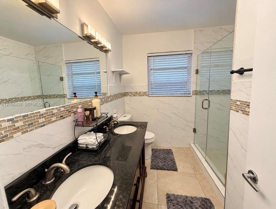 Bathroom featuring a dual-sink vanity with a dark stone countertop, a glass-enclosed walk-in shower with stone tile wall accents, and large-format tile flooring