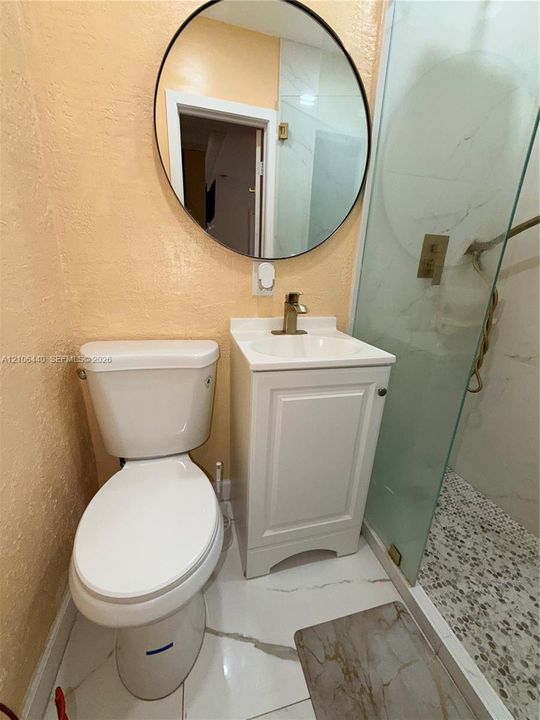 Compact bathroom featuring a white single-sink vanity with metallic hardware, a round wall-mounted mirror, a walk-in shower with stone-style floor tiling, and large-format tile flooring