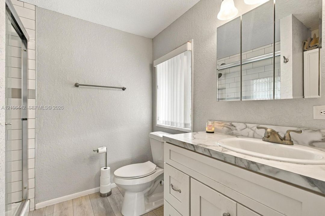 Bathroom featuring a white vanity with a patterned stone-finish countertop, integrated sink, three-panel medicine cabinet, tiled walk-in shower, and wood-finish flooring
