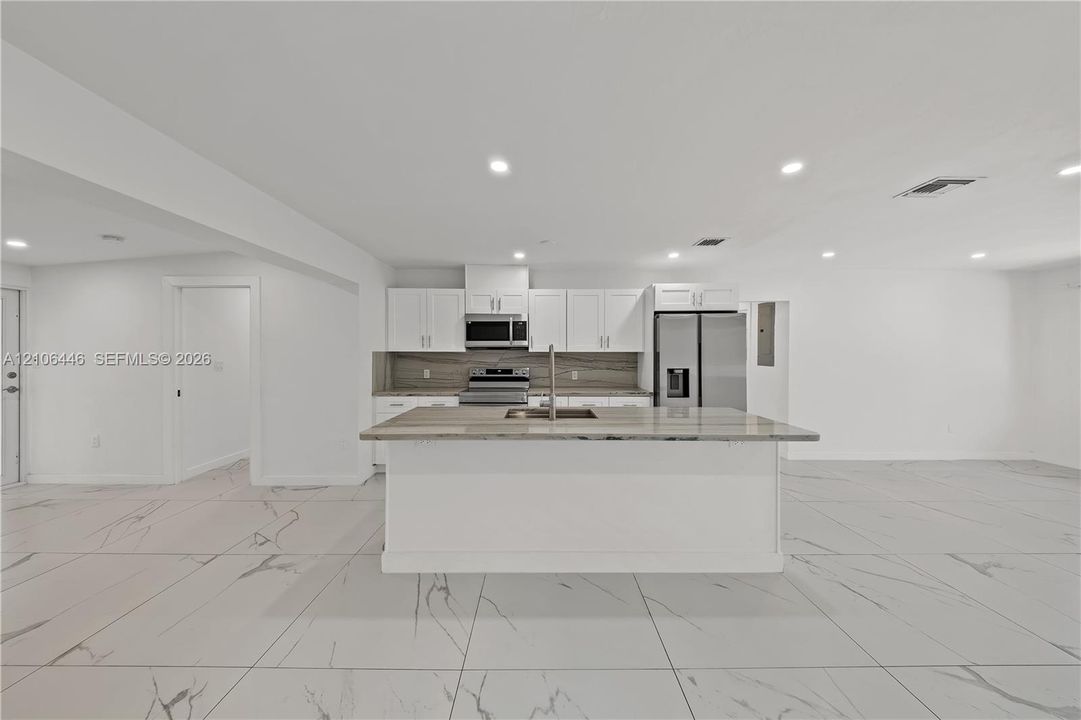 Kitchen featuring white shaker-style cabinetry, stone-patterned countertops, a central island with an integrated stainless steel sink, stainless steel appliances, and large-format tile flooring with a veined pattern