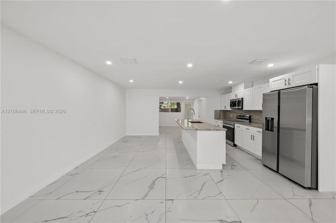 Open-concept kitchen featuring large-format tile flooring, white cabinetry, stone countertops, a center island with an integrated sink, and stainless steel appliances