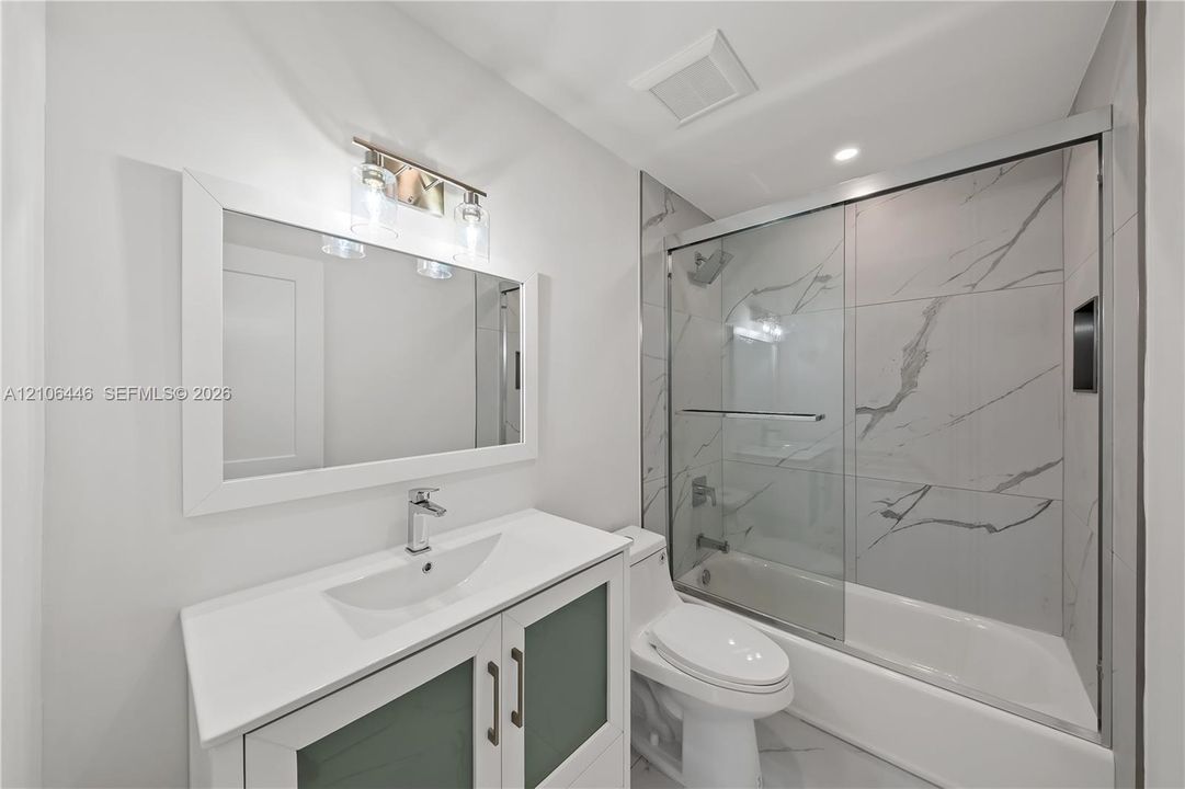 Bathroom featuring a vanity with a solid-surface countertop and integrated basin, large framed mirror, vanity lighting, and a bathtub with glass sliding doors and full-height stone-finish wall tile