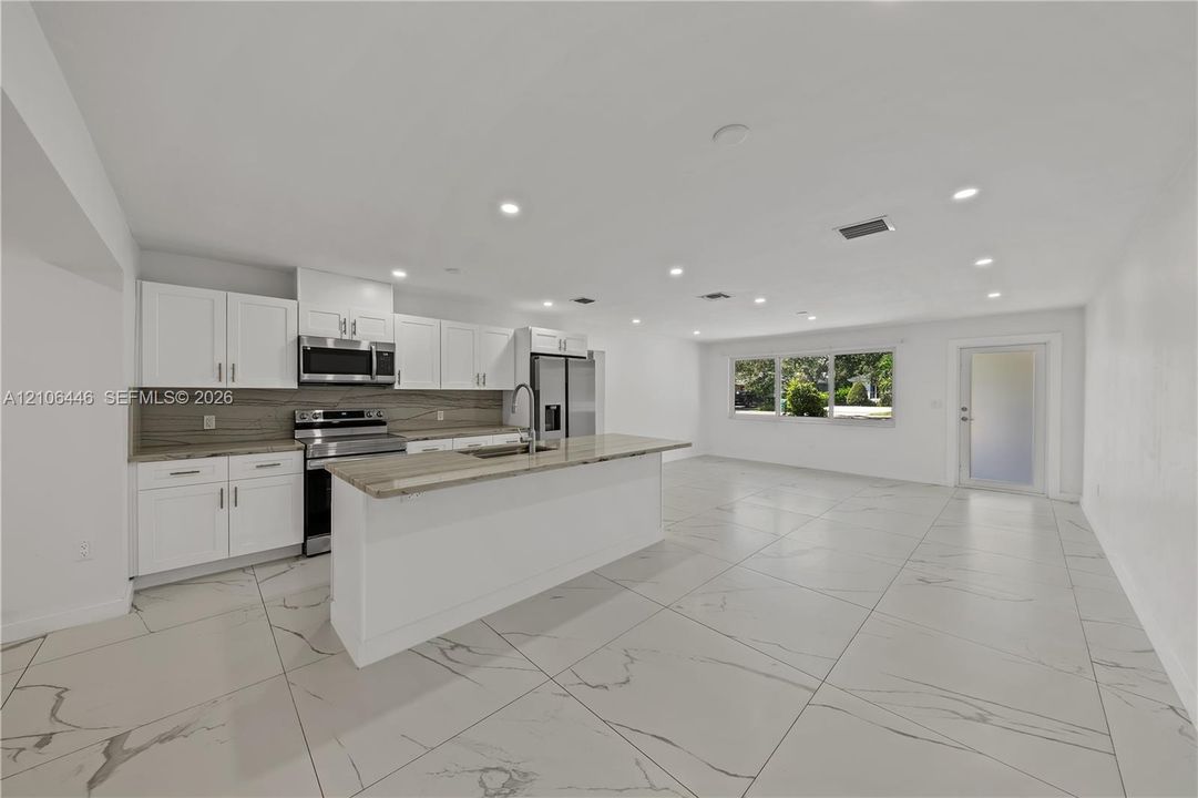 Kitchen featuring white shaker cabinetry, stone-finish countertops, stainless steel appliances, a central island with an integrated sink, and large-format porcelain tile flooring
