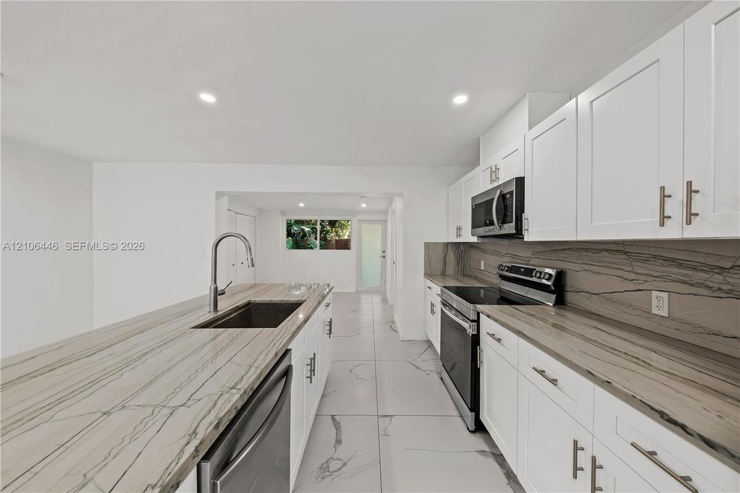 Kitchen featuring white shaker-style cabinetry, natural stone-patterned countertops, a full-height stone backsplash, large-format tile flooring, and stainless steel appliances