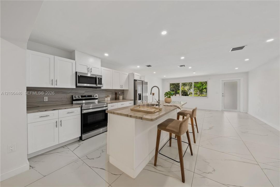 Kitchen featuring white cabinetry, stone-finish countertops, stainless steel appliances, a center island with a sink, and large-format tile flooring