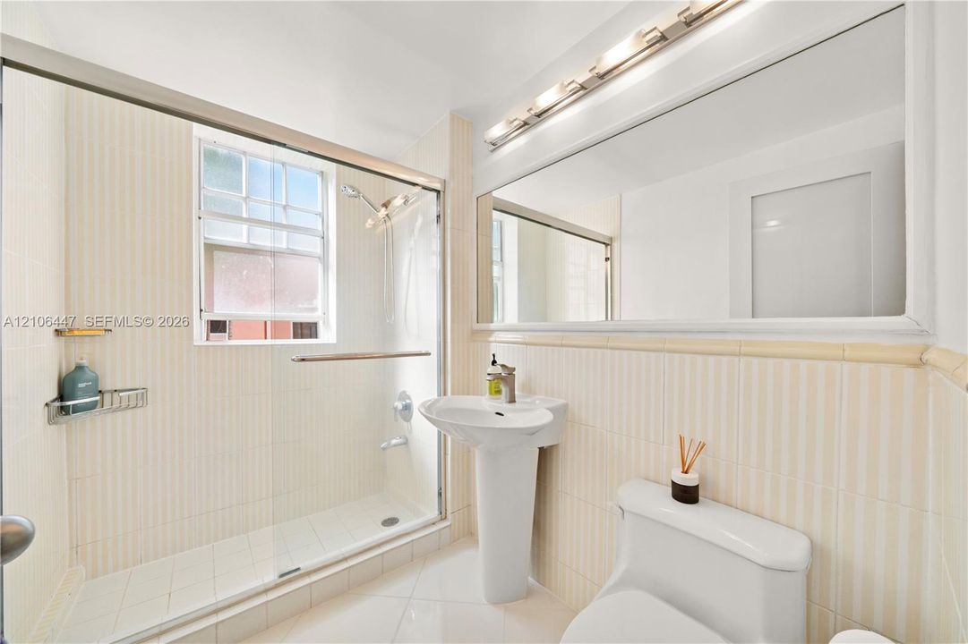 Full-width vanity mirror with integrated multi-bulb lighting, pedestal sink, glass-enclosed walk-in shower with tiled surround, and ceramic tile flooring