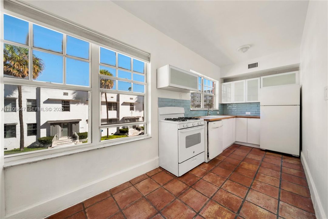 Kitchen featuring large multi-pane windows, terracotta tile flooring, white cabinetry with glass-front uppers, and blue tile backsplash