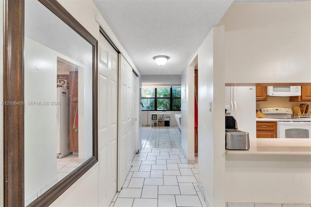 Hallway featuring large-format ceramic tile flooring, double-door closet cabinetry, ceiling-mounted light fixture, and a pass-through kitchen opening with a countertop ledge