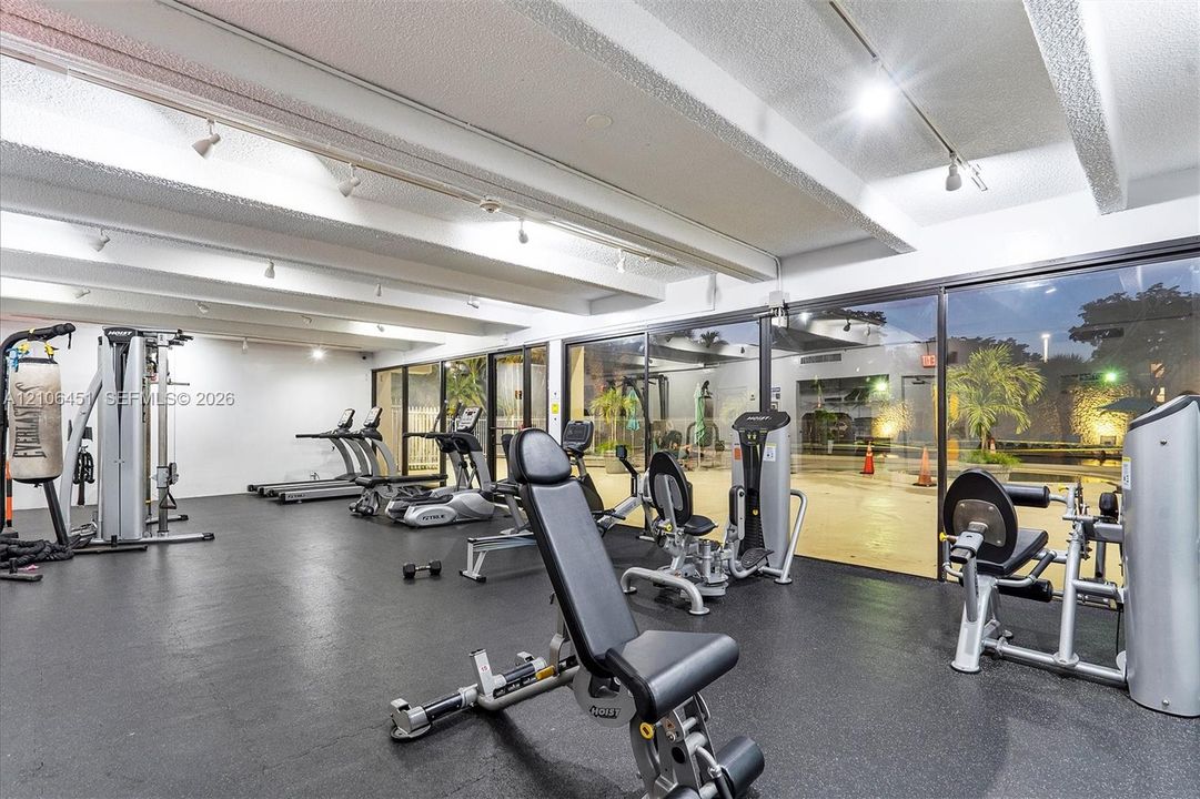 Expansive fitness center featuring rubberized flooring, exposed beam ceiling, adjustable track lighting, and floor-to-ceiling glass wall panels