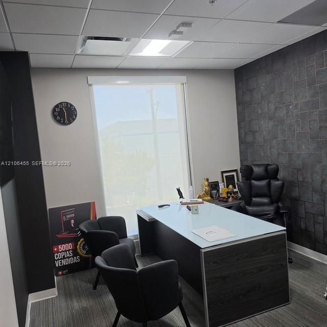 Office space featuring a textured dark stone accent wall, neutral wall tones, grid-pattern ceiling tiles, and window with a roller shade