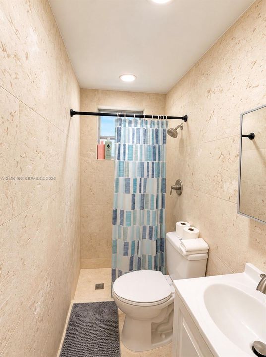 Full-height wall-to-wall stone tile, built-in vanity with integrated basin, wall-mounted medicine cabinet, recessed lighting, and shower area with matte black hardware