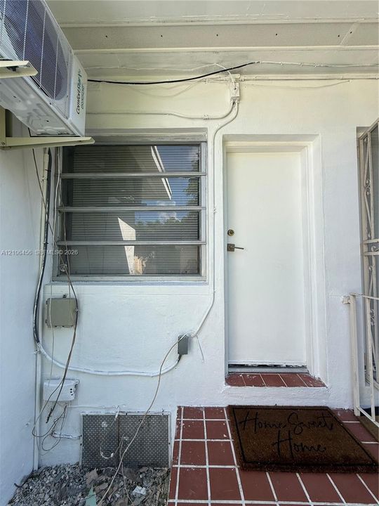 Exterior entryway featuring a solid-core door, tiled landing, jalousie window with security bars, and a wall-mounted climate control unit
