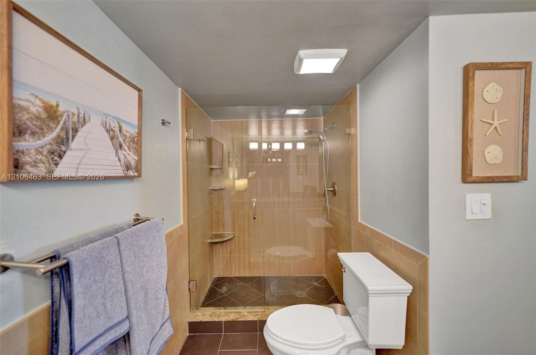 Walk-in glass enclosed shower with neutral wall tiling, stone floor tile, built-in corner shelf, and wall-mounted hand shower fixture