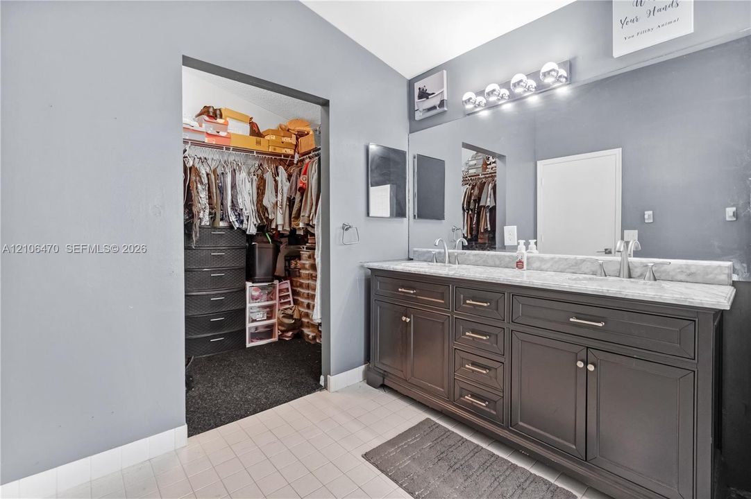 Dual-vanity cabinet with dark finish and light-toned stone countertop, large-format wall mirror, multi-bulb vanity light fixture, and built-in closet storage