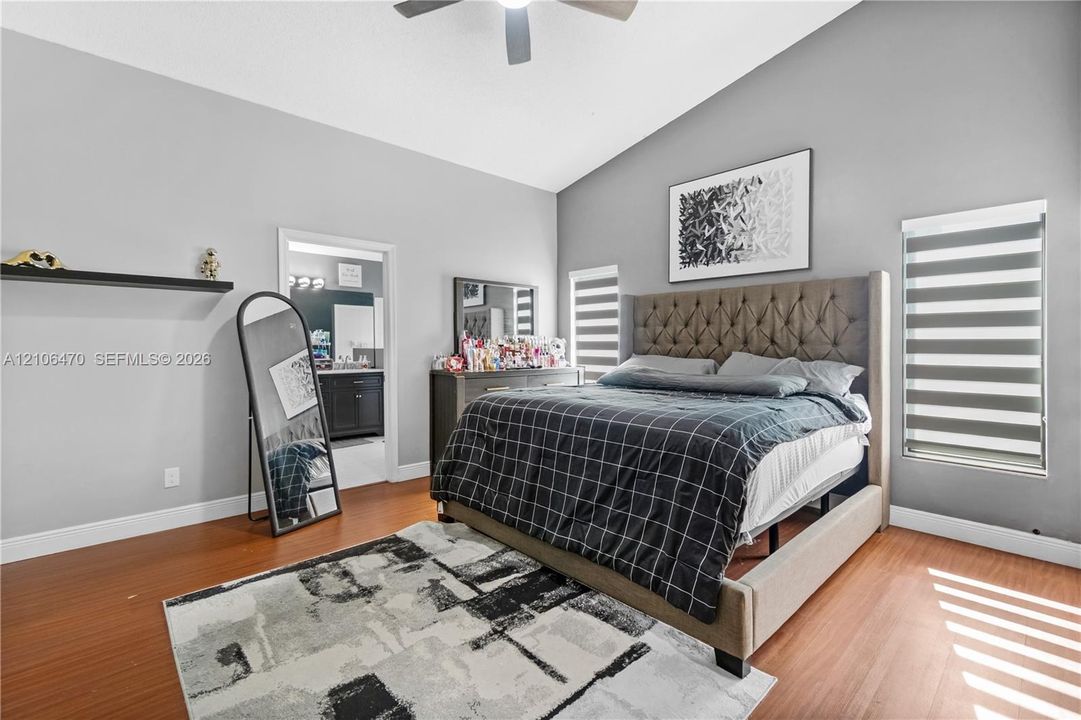 Vaulted ceiling with a centrally mounted ceiling fan, wood-finish flooring, two windows featuring dual-layer roller shades, and a built-in wall-mounted floating shelf