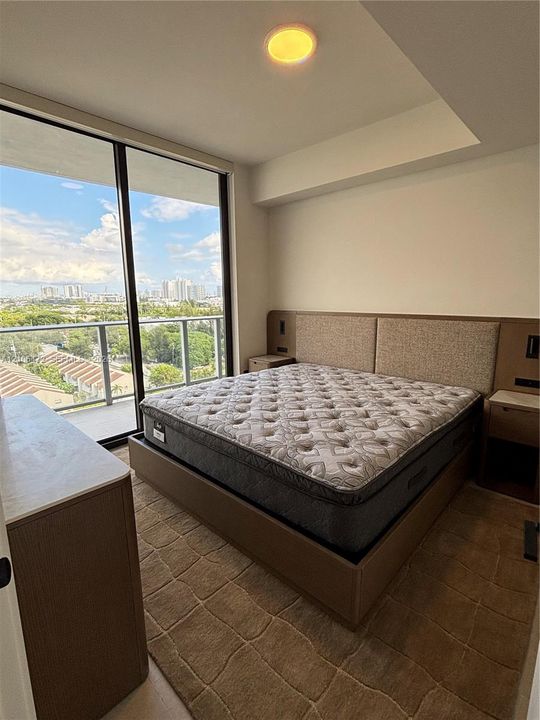 Floor-to-ceiling glass sliding doors, private balcony with metal railing, built-in wooden headboard with integrated lighting and electrical outlets, recessed ceiling lighting, and solid-surface topped wooden cabinetry
