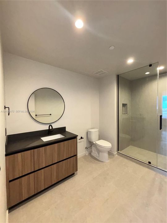 Bathroom featuring a wood-finish floating vanity with a dark countertop and integrated sink, circular wall-mounted mirror, matte black faucet, and a walk-in glass shower enclosure