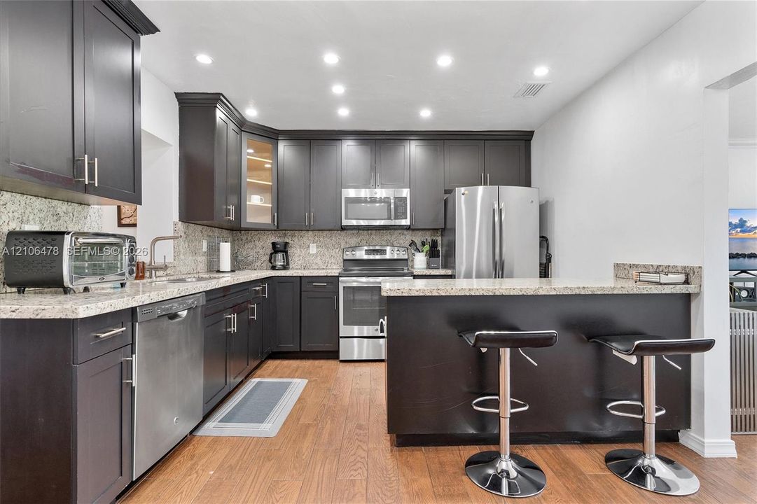 Kitchen featuring dark cabinetry, stone-surface countertops, wood-finish flooring, stainless steel appliances, and a peninsula with seating