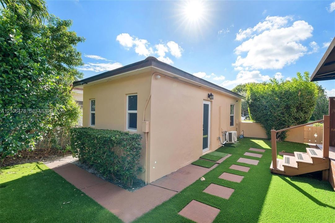 Detached structure with stucco exterior, synthetic turf lawn, stone paver pathway, and wood deck stairs