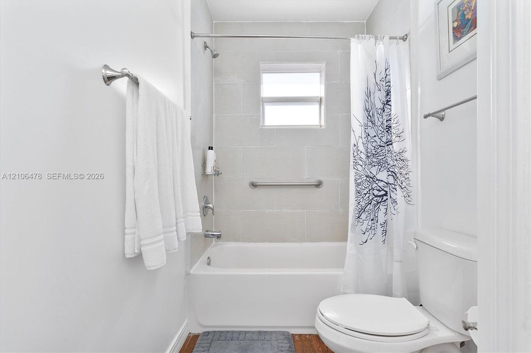 Full bathroom featuring a white porcelain tub, light-toned stone-finish tile shower surround, integrated grab bar, single-handle shower faucet, and wall-mounted metal towel hardware