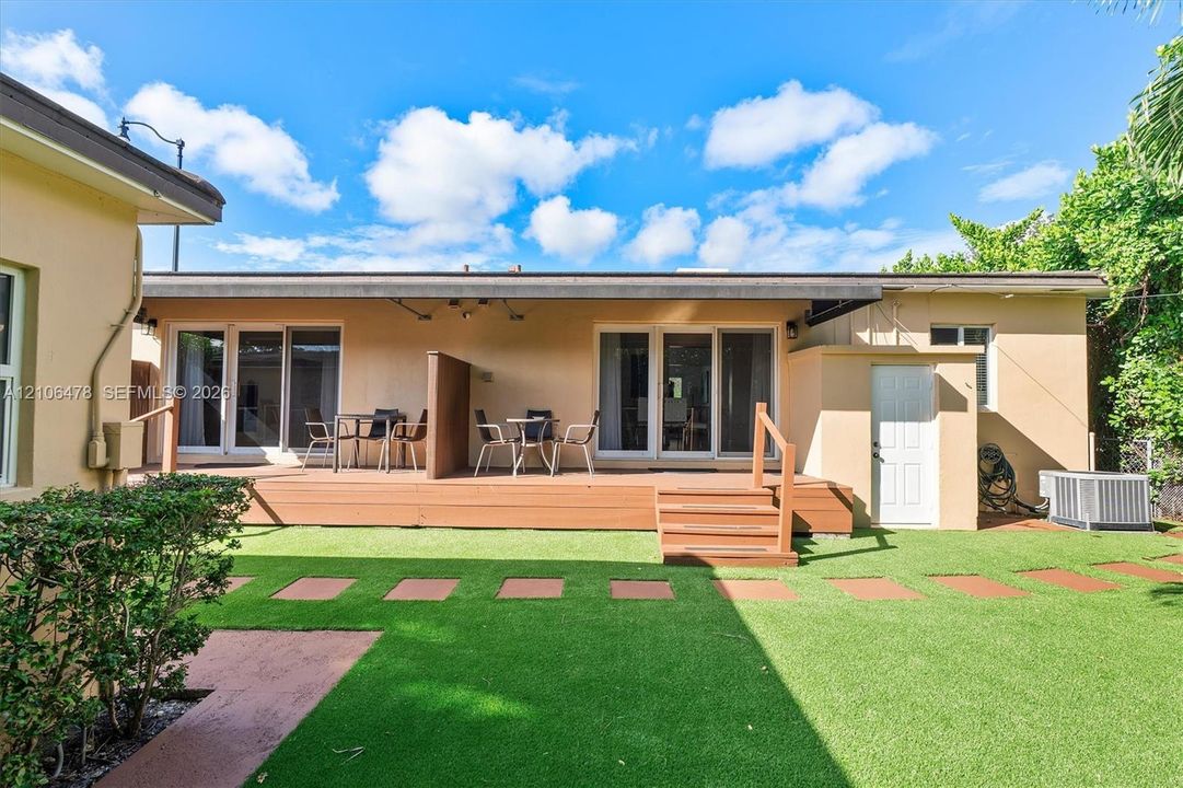 Backyard featuring a raised composite deck, synthetic lawn, square stone pavers, and multiple sliding glass door entries