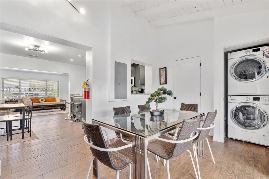 Open-concept interior featuring wood-finish flooring, vaulted white-painted wood beamed ceiling, pass-through window to kitchen, and built-in stacked laundry unit