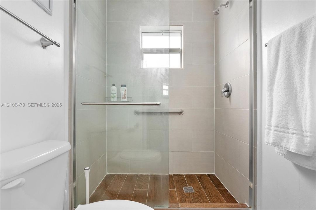 Walk-in shower featuring light-toned wall tile, wood-finish floor tile, a glass door enclosure, and a built-in metal grab bar