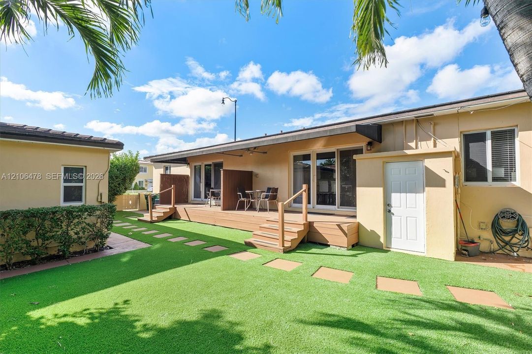Backyard area featuring a raised wood-finish deck, synthetic turf lawn, stone paver pathway, and dual sliding glass door entryways