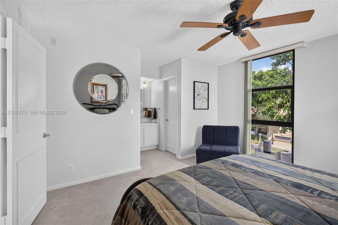 Neutral wall-to-wall carpeting, multi-panel interior door, ceiling fan with wood-finish blades, and large fixed-pane window with black framing